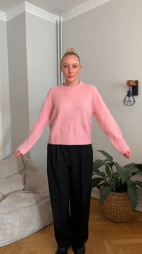 Svetlo roza ohlapni pulover z rebrastim robom in manšetami, kombiniran s črnimi, širokimi hlačami. V ozadju tkan cvetlični lonec.