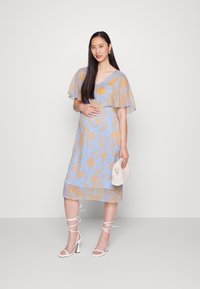 Vestido floral em azul claro com flores amarelas, mangas curtas e soltas, decote em V e até ao joelho. Sandálias brancas de salto alto e uma pequena mala.