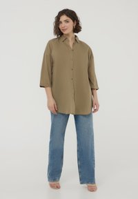 Camicia khaki con bottoni, dal taglio morbido e maniche a tre quarti, abbinata a jeans dritti in denim chiaro. Tessuto morbido al tatto.