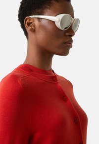 Sonnenbrille mit weißen Rahmen und runden, reflektierenden Gläsern. Das Modell trägt eine rote Strickjacke mit Knopfdetails in der Nähe des Halsausschnitts.