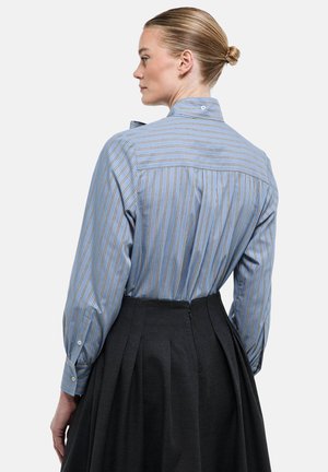 Femme aux cheveux blonds attachés en chignon, portant une chemise à manches longues rayée bleue rentrée dans une jupe plissée foncée taille haute, vue de profil.