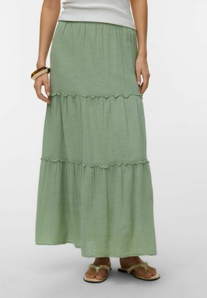 Žena nosí dlhú, svetlozelenú vrstvenú sukňu v kombinácii s bielymi sandálmi a bielym topom, stojac pred jednoduchým pozadím.