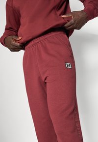 Bordeaux farvede sweatpants med elastisk talje, der har sidelommer og et lille sort-hvidt logo på fronten. Glat, struktureret stof.