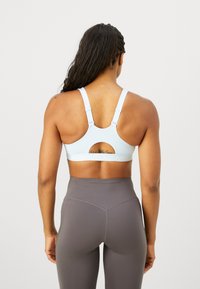 Sostén deportivo azul claro con un corte redondo en la parte posterior, tirantes ajustables y una banda texturizada con el logo de Nike. Combinado con leggins gris oscuro.