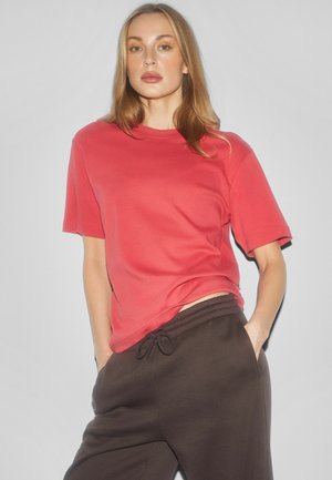 Kobieta o jasnych włosach, ubrana w koralowy T-shirt z krótkim rękawem i ciemnobrązowe spodnie z troczkiem, stoi z rękami w kieszeniach na jednolitym tle.