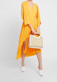 Keltainen laskostettu mekko, jossa on V-pääntie ja pitkät hihat, yhdistettynä rakenteelliseen laukkuun, jossa on olkikuitutekstuuri ja valkoiset korostukset. Valkoiset mulesit jalassa.