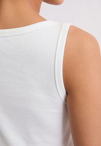Weißes geripptes Tanktop mit runder Ausschnittlinie, das über eine glatte Textur und eine figurbetonte Passform verfügt und die Schulter sowie einen Teil des Arms zeigt.