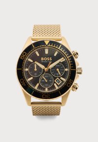 Χρυσόχρωμο ρολόι χειρός Hugo Boss με μαύρο καντράν, τρεις υποκαντράν, παράθυρο ημερομηνίας στη θέση 4 και μπρασελέ από δίχτυ.