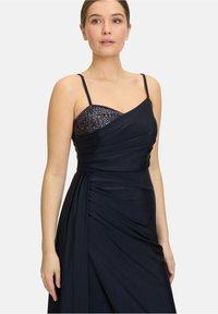 Robe de soirée marine à fines bretelles, avec un corsage perlé et des détails drapés. Tissu épuré avec une texture lisse.