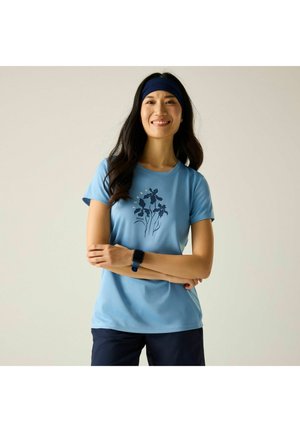 Kobieta z długimi czarnymi włosami ubrana w niebieski kwiecisty t-shirt, granatowe spodnie, czarny smartwatch i granatową opaskę na głowę, uśmiechająca się ze skrzyżowanymi rękami.