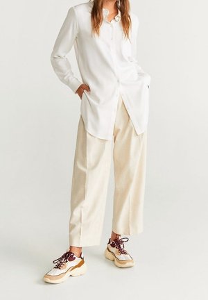 Woman wearing oversized white button-up shirt, loose cream-colored pants, and chunky white sneakers with maroon laces, standing with hands in pockets.