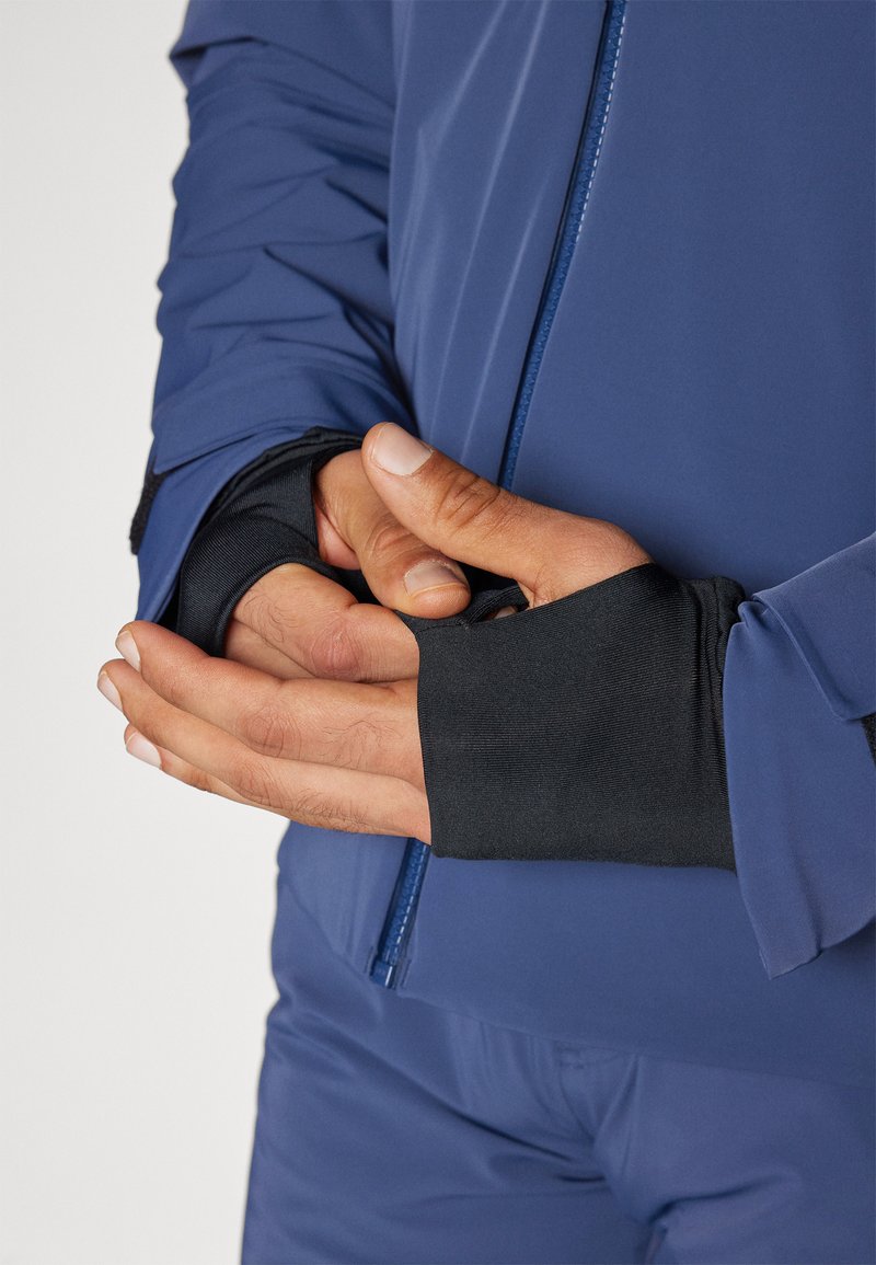 Veste de sport bleue avec des poignets noirs, présentant une fermeture éclair et une coupe ajustée. Texture lisse et ajustement serré au niveau des poignets.