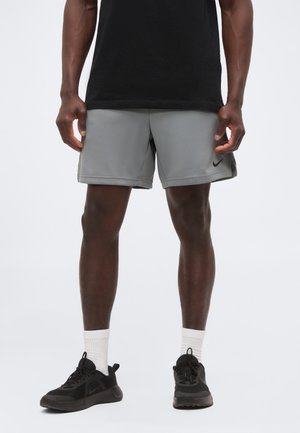 Άνδρας που φοράει γκρι αθλητικό σορτς, μαύρα αθλητικά παπούτσια με λευκές κάλτσες και μαύρο πουκάμισο, στέκεται μπροστά σε ένα απλό φωτεινό φόντο.