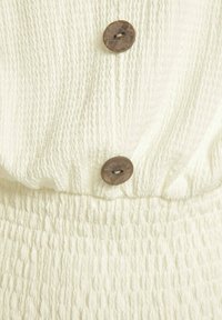 Vit texturerad tygblus med smockad midja och två mörka runda knappar fram, med ett rynkat mönster och mjuk känsla.