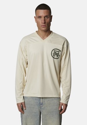 Mężczyzna w beżowej siateczkowej koszuli z długim rękawem z ciemnym logo na lewej piersi, ubrany w jasnoniebieskie dżinsy, stoi na białym tle.