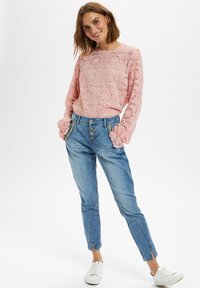 Top lungo a maniche lunghe in pizzo rosa con dettagli di pattern testurizzati, abbinato a jeans tapered azzurri chiari con tasche laterali e dettagli con bottoni. Sneakers bianche.