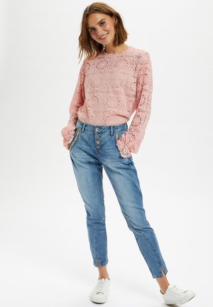 Haut à manches longues en dentelle rose avec des détails de motif texturé, associé à un jean tapered bleu clair avec des poches latérales et des accents de boutons. Baskets blanches.