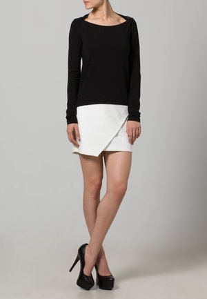 Kvinde iført en sort langærmet top, hvid asymmetrisk miniskørt og sorte højhælede platformssko, stående med benene krydset.