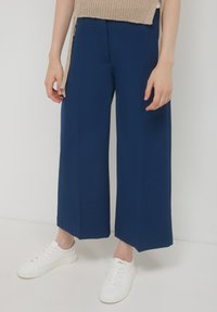 Persona che indossa pantaloni a palazzo blu navy e sneakers bianche, in piedi contro uno sfondo chiaro e uniforme.