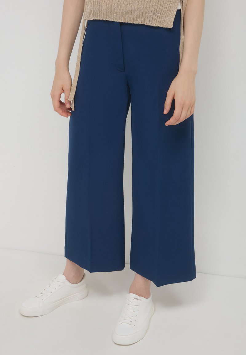 Persona che indossa pantaloni a palazzo blu navy e sneakers bianche, in piedi contro uno sfondo chiaro e uniforme.