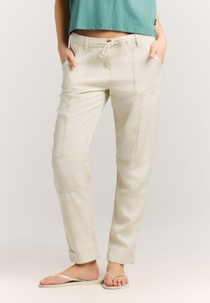 Osoba oblečená v béžových nohaviciach so šnúrkou na stiahnutie a ohrnutými lemy, nosiaca žabky, ruky vreckách, stojaca pred jednoliatym pozadím.