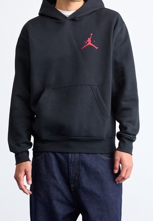 Man wearing black hooded sweatshirt with front pocket and small red Jumpman logo on left chest, paired with dark blue jeans.