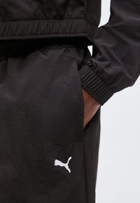 Χέρι στην τσέπη φοράει μαύρο παντελόνι Puma με ελαστικό τελείωμα και ορατό λευκό λογότυπο Puma στο πόδι.