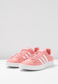 Un paio di sneaker basse in suede rosa con righe e suole bianche, posizionate su una superficie bianca contro uno sfondo grigio chiaro.