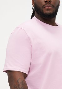 Tricou din bumbac roz deschis cu guler rotund și mâneci scurte. Prezintă o textură netedă și un design simplu, arătând parțial un tatuaj pe braț.