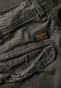Un pantalon utilitaire noir avec un tissu texturé et des coutures visibles. Une étiquette portant la mention "SUPERDRY" est cousue sur la poche arrière.