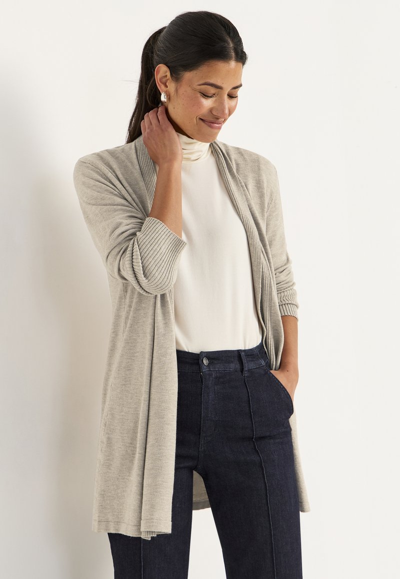 Vrouw met donker haar in een paardenstaart draagt zilveren oorbellen, een beige cardigan over een witte coltrui en donkerblauwe broek, staand voor een effen achtergrond.