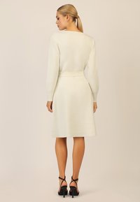 Weißes gestricktes Kleid mit langen Ärmeln, tailliertem Schnitt und ausgestelltem Rock. Kombiniert mit schwarzen Riemchen-High Heels. Glatte Textur.
