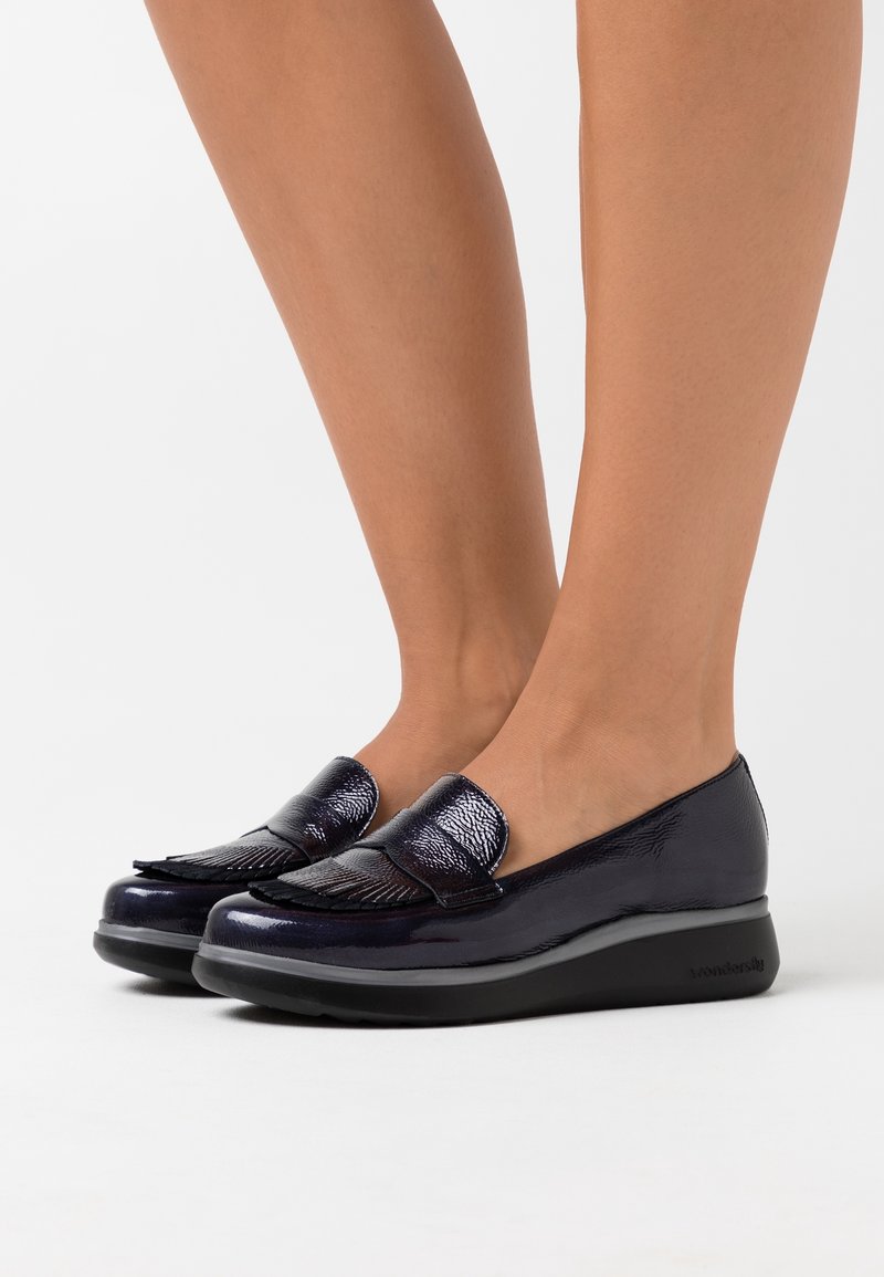 Mocassins en cuir noir brillant avec détail de franges portés pieds nus, vus de côté sur fond blanc.