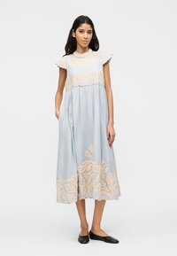 Γυναίκα που στέκεται, φορώντας ένα γαλάζιο μίντι φόρεμα με μπεζ λουλουδένιο κέντημα, κοντά μανίκια και μαύρα επίπεδα παπούτσια σε απλό φόντο.