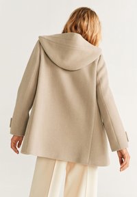 Manteau en laine beige avec capuche, présentant une coupe décontractée, des manches longues et des poignets boutonnés. Texture lisse et silhouette en A.