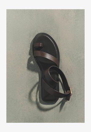 Sort flad sandal med to tynde mørkebrune remme over tæerne og en bred ankelrem med guldlås på grå overflade.