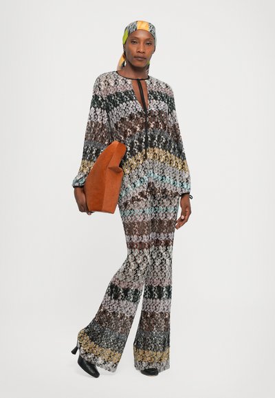 Woman wearing multicolored patterned knit top and pants with black heels, colorful headscarf, holding large rust-orange bag.