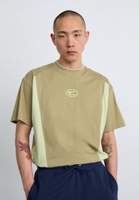 Béžové oversized tričko s svetlozelenými bočnými panelmi a malým logom Nike. Vyrobené z mäkkého materiálu, s okrúhlym výstrihom a krátkymi rukávmi.