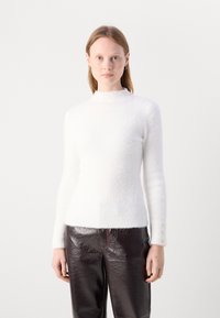 Weißer, strukturierter Pullover mit hohem Kragen und langen Ärmeln, die am Bündchen mit drei Knöpfen versehen sind. Kombiniert mit glänzenden, dunkelbraunen Hosen.