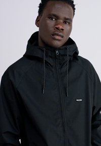 Chaqueta negra con cremallera y capucha, con cordones en contraste, un logo en el pecho y una textura suave.