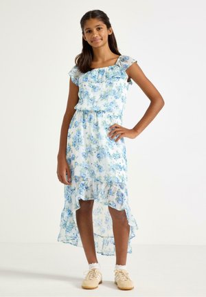 Tyttö seisoo käsi lanteilla valkoisessa ja sinisessä kukallisessa liehuvassa korkeavyötäröisessä mekossa sekä beigenvärisissä lenkkareissa, joissa on valkoiset nilkkasukat.