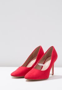 Czerwone zamszowe szpilki z wydłużonym noskiem, gładką fakturą i smukłym obcasem typu stiletto. Wnętrze ma jasnobrązowe wykończenie.