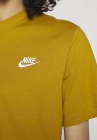 Žluté bavlněné tričko s bílým vyšitým logem Nike na levé části hrudi, s klasickým kulatým výstřihem a krátkými rukávy.
