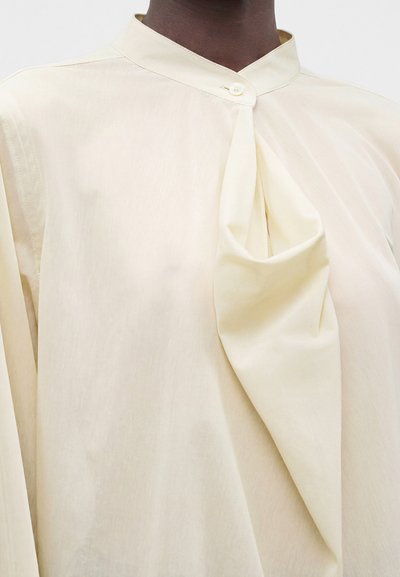 Gros plan d’une personne portant un chemisier couleur crème avec un col montant et un détail unique en tissu drapé sur le devant.