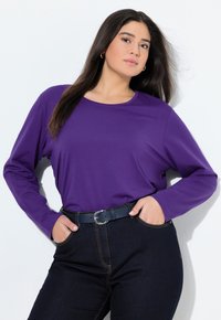 Paarse lange mouwtop met ronde hals, gecombineerd met donkerblauwe hoogtaille jeans. Het model heeft de handen in de zakken en draagt een zwarte riem. Soepele stof.