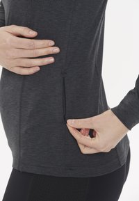Tummanharmaa pitkähihainen paita, jossa on sivutasku ja vetoketjuyksityiskohta. Valmistettu pehmeästä kankaasta, jossa on vartalonmyötäinen muotoilu ja litteät saumat.