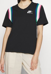 Černé sportovní tričko Nike s výstřihem do V a růžovými, bílými, zelenými a modrými pruhy na ramenou, nošené s světle béžovými kalhotami.