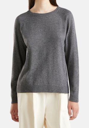 Frau trägt einen eng anliegenden, langärmligen grauen Strickpullover und lockere, cremefarbene Hosen, steht vor einem einfarbigen Hintergrund.