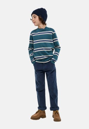 Chemise à manches longues rayée en bleu sarcelle avec des rayures blanches et crème, associée à un pantalon bleu marine et des bottes à lacets marron.