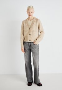 Μπεζ μάλλινη κάρντιγαν με ριμπ άκρες, δύο μπροστινές τσέπες και πέντε κουμπιά, συνδυασμένη με γκρι φαρδιά παντελόν�ια και σκούρα παπούτσια.